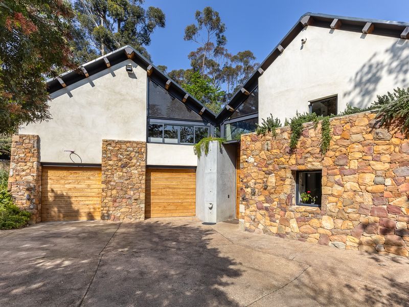 Modern architectural home exterior featuring a combination of natural stone cladding, concrete elements, and timber garage doors with a paved driveway.