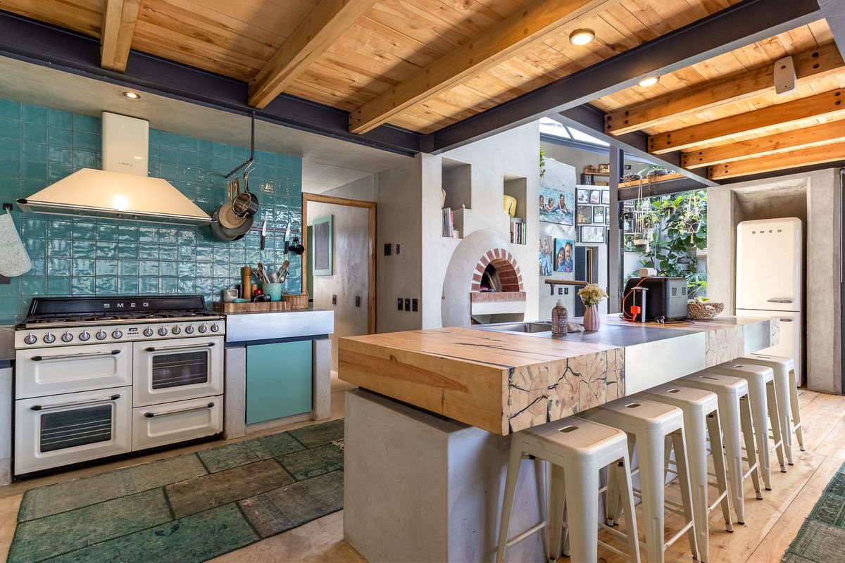 A stylish kitchen featuring exposed timber and steel beams, a large concrete island with a timber top, a built-in pizza oven, and a gas range.