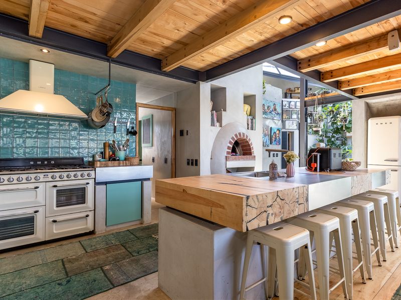 A stylish kitchen featuring exposed timber and steel beams, a large concrete island with a timber top, a built-in pizza oven, and a gas range.