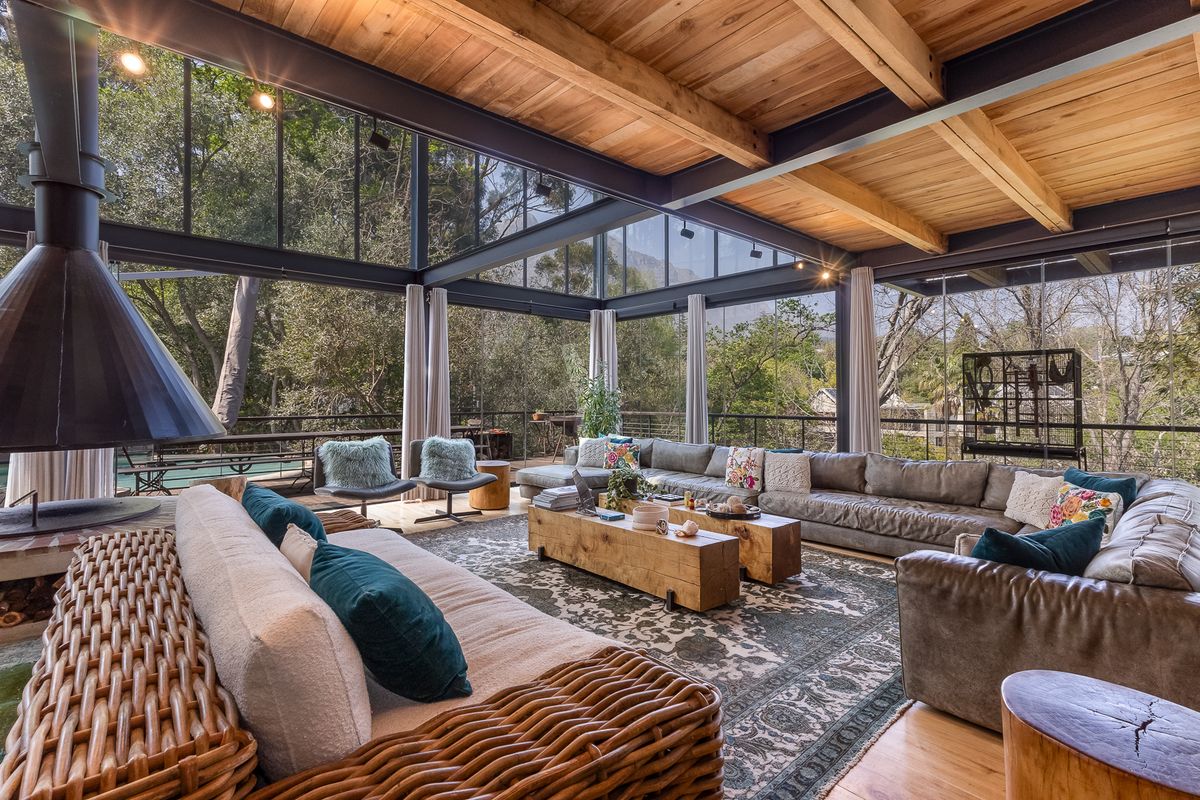 Spacious living area featuring a striking suspended fireplace, exposed timber beams, and expansive floor-to-ceiling glass walls offering panoramic views of the surrounding greenery.