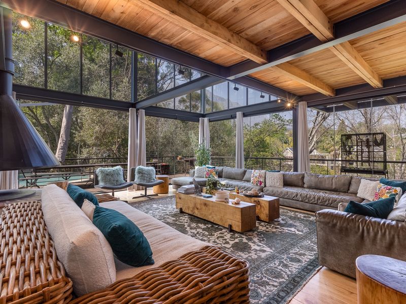 Spacious living area featuring a striking suspended fireplace, exposed timber beams, and expansive floor-to-ceiling glass walls offering panoramic views of the surrounding greenery.