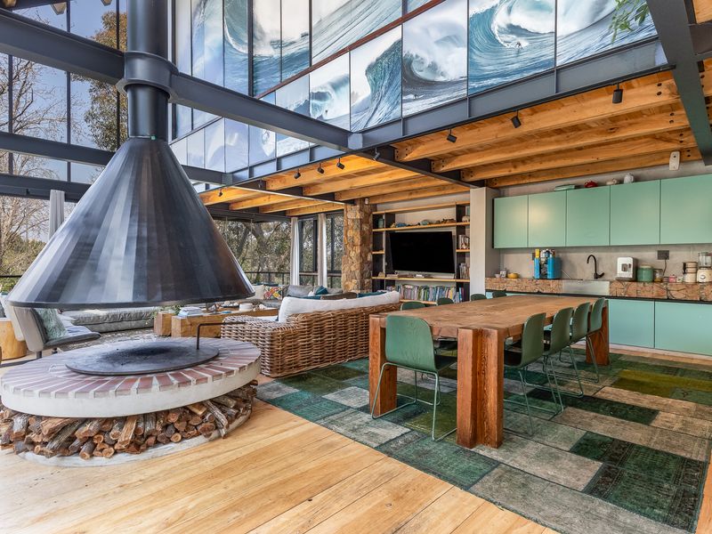 Spacious open-plan living and dining area featuring a striking suspended fireplace, exposed timber beams, and modern kitchen cabinetry with expansive glass walls.