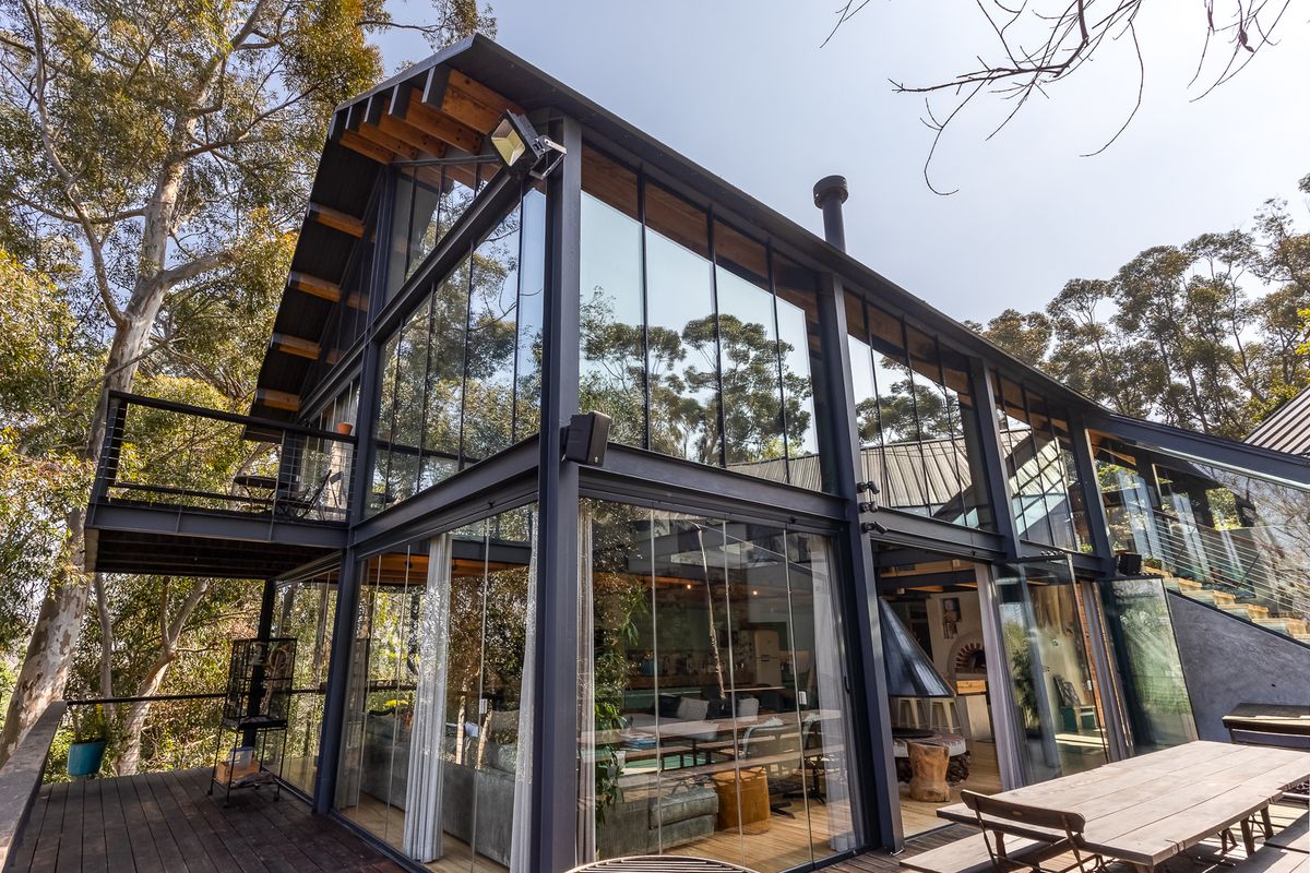 Modern architectural home featuring extensive steel framing, floor-to-ceiling glass walls, and expansive timber decking nestled in a natural wooded setting.