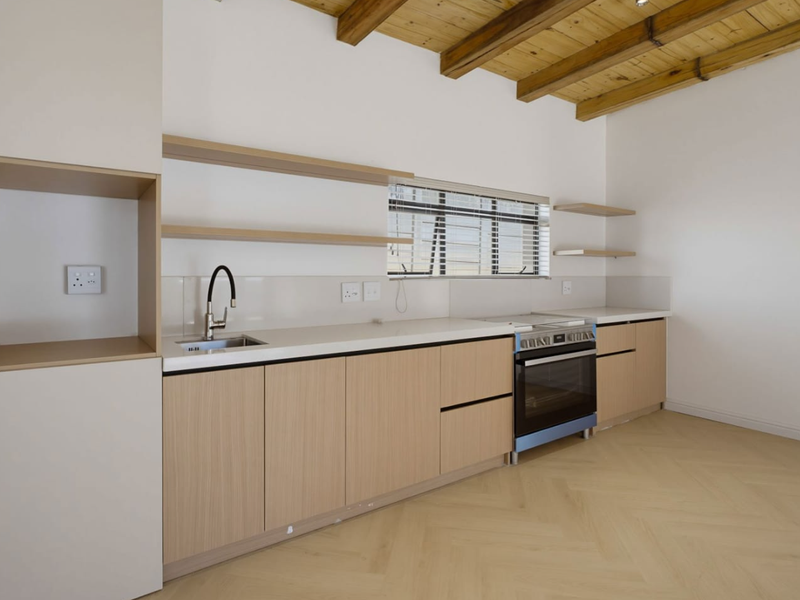 Modern kitchen featuring light wood cabinetry, stone countertops, exposed timber ceiling beams, and herringbone flooring in a bright, open-plan layout.