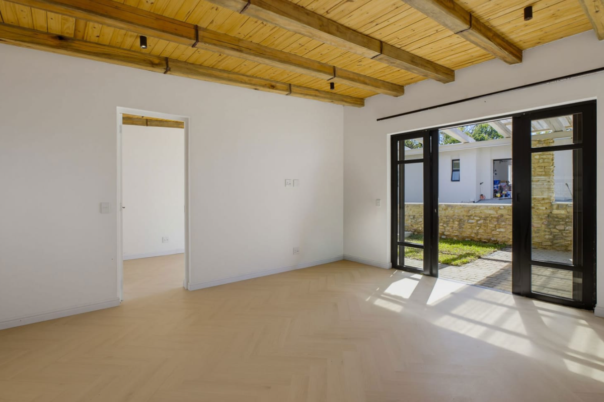 Bright, empty living area featuring exposed timber ceiling beams, modern herringbone flooring, and large black aluminium sliding doors opening to a paved patio.