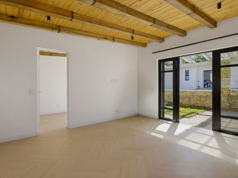 Bright, empty living area featuring exposed timber ceiling beams, modern herringbone flooring, and large black aluminium sliding doors opening to a paved patio.