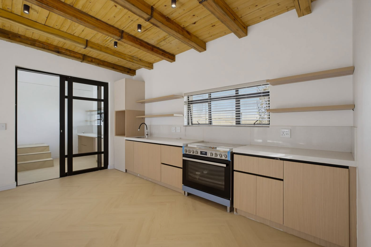 Modern kitchen featuring light wood cabinetry, stone countertops, built-in oven, and exposed timber beam ceiling, leading to an adjacent room through a black framed door.