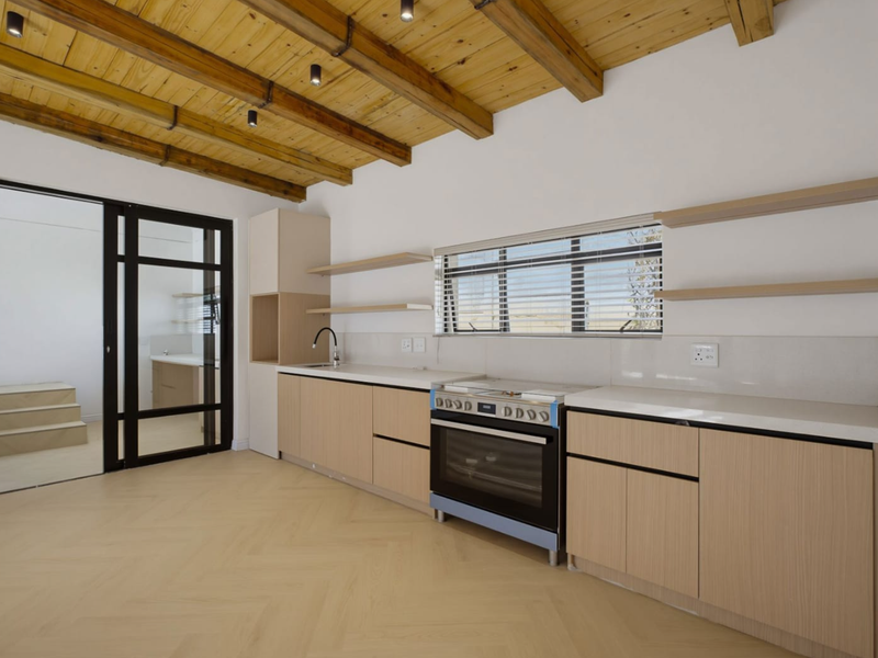 Modern kitchen featuring light wood cabinetry, stone countertops, built-in oven, and exposed timber beam ceiling, leading to an adjacent room through a black framed door.