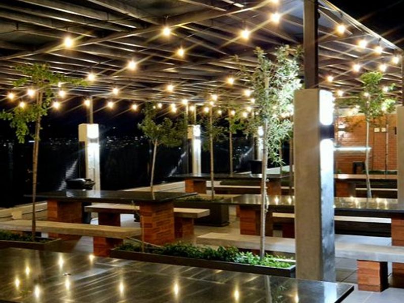 A spacious covered outdoor patio featuring granite-topped tables with brick bases, integrated planters, and ambient string lighting, ideal for evening entertainment.