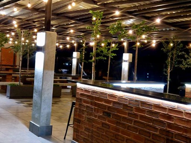 A well-lit covered outdoor patio featuring an exposed brick bar counter, tiled flooring, and modern structural lighting, ideal for evening entertainment.
