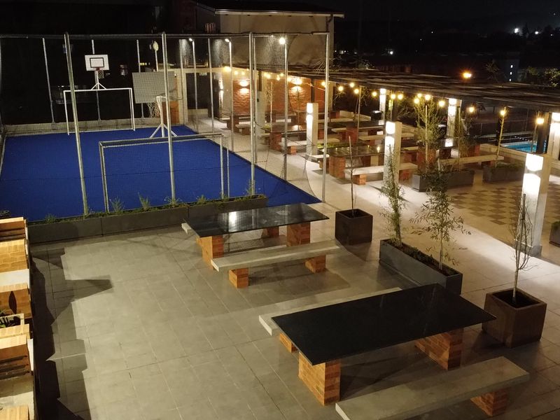 A well-maintained rooftop sports facility featuring a fenced court, integrated seating areas with stone tables, and ambient overhead lighting.