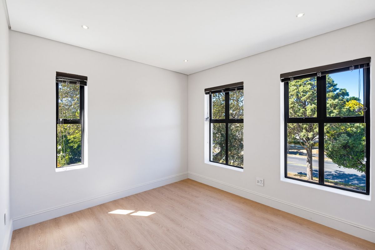 A bright, empty bedroom featuring modern laminate flooring, recessed downlights, and large black-framed windows with venetian blinds.
