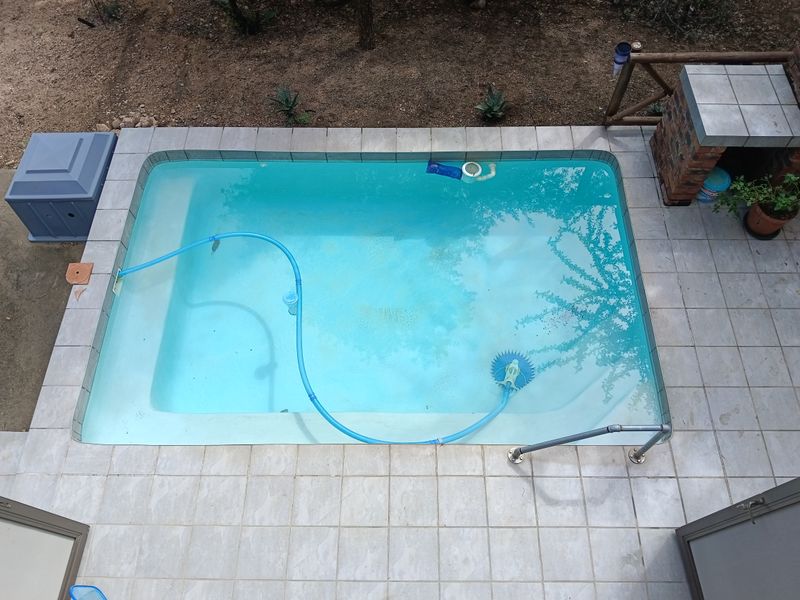 An elevated view of a rectangular swimming pool featuring a tiled surround, a built-in brick braai area, and a metal handrail.