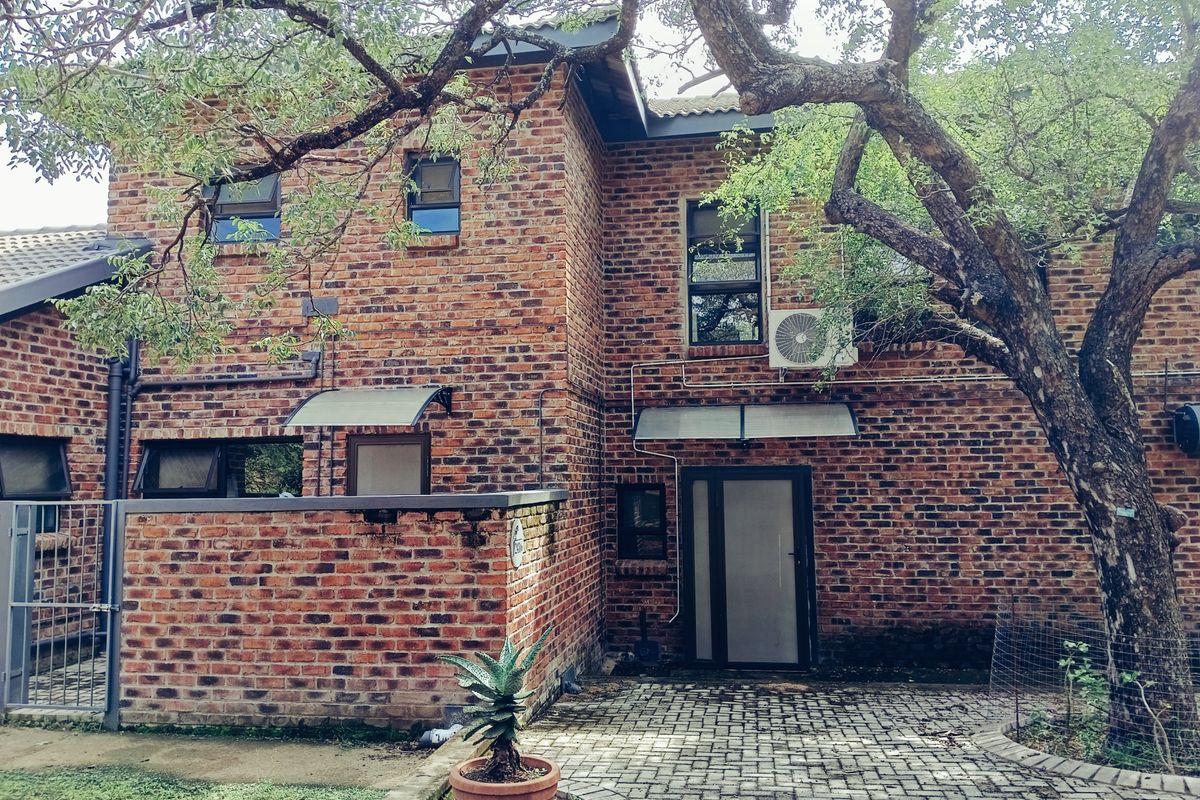 Rear view of a double-story face brick house featuring a paved patio area, mature trees, and an external air conditioning unit.