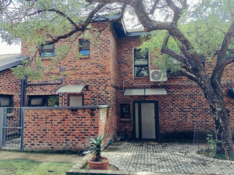 Rear view of a double-story face brick house featuring a paved patio area, mature trees, and an external air conditioning unit.