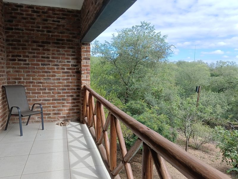 A covered balcony featuring exposed brickwork, tiled flooring, and a wooden balustrade, offering a peaceful view of the surrounding natural greenery.