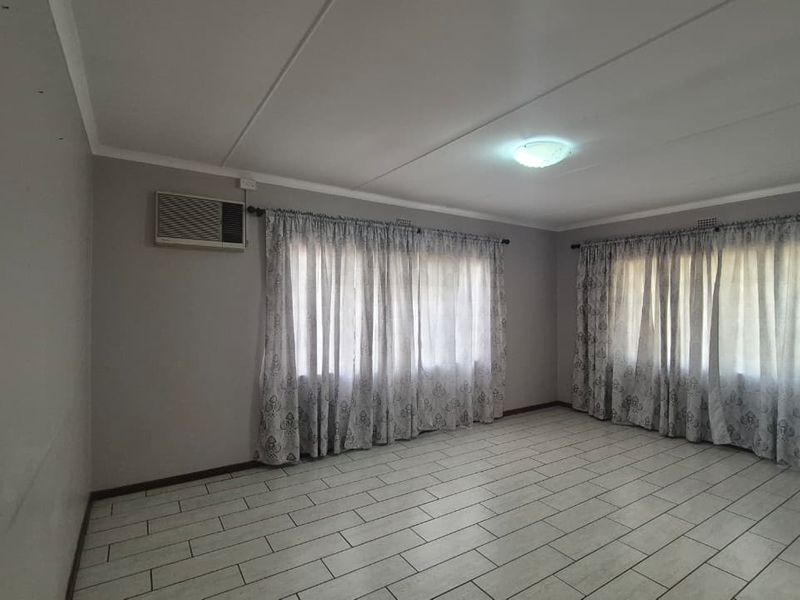 A spacious, empty living room featuring neutral tiled flooring, a wall-mounted air conditioning unit, and large windows with curtains.