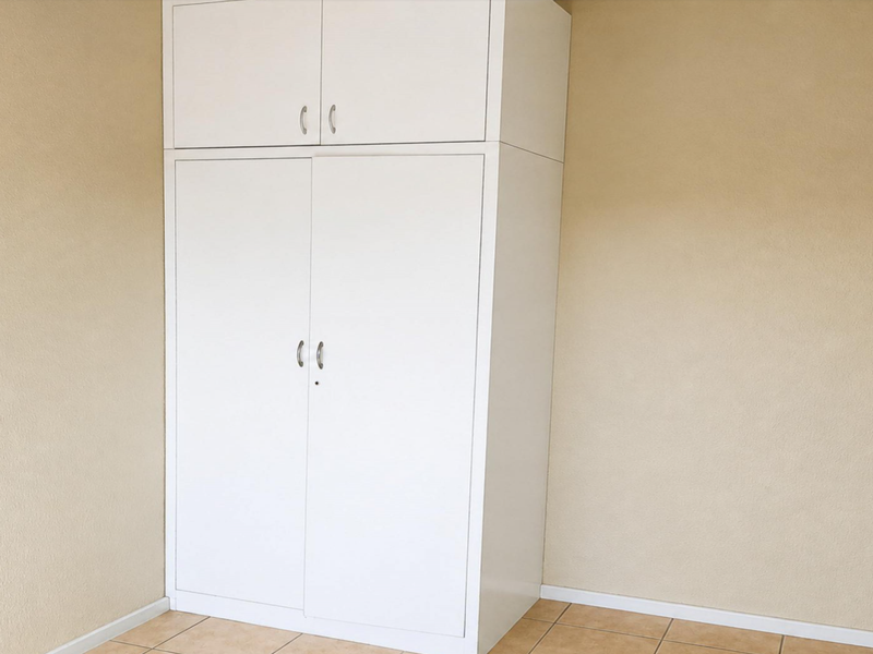 A clean, empty bedroom featuring neutral wall tones, tiled flooring, and a white built-in wardrobe with overhead storage.