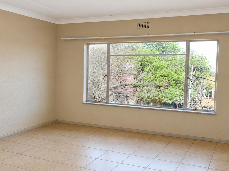 A spacious, empty bedroom featuring neutral tiled flooring, a large window with a curtain rail, and simple cove cornicing.