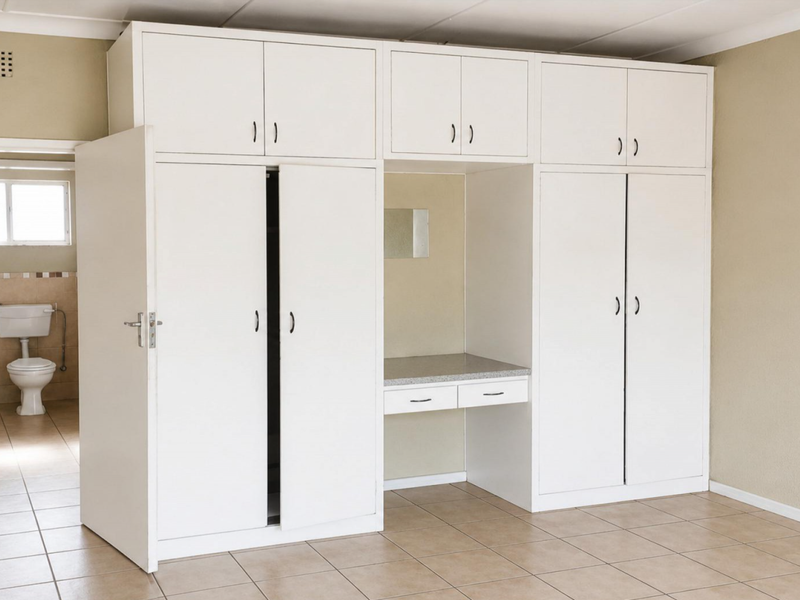 A spacious bedroom featuring extensive white built-in cupboards with an integrated vanity desk, tiled flooring, and direct access to an en-suite bathroom.