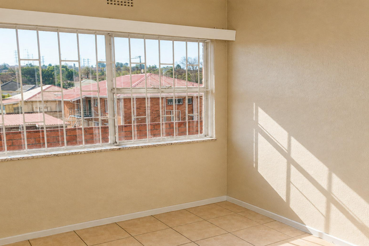 An empty bedroom featuring tiled flooring and a large window with security bars, offering a view of the surrounding neighborhood.