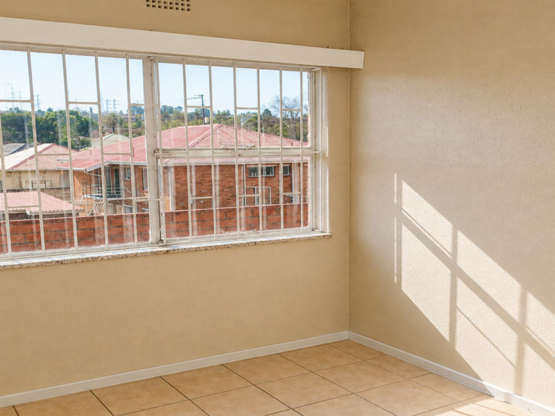An empty bedroom featuring tiled flooring and a large window with security bars, offering a view of the surrounding neighborhood.