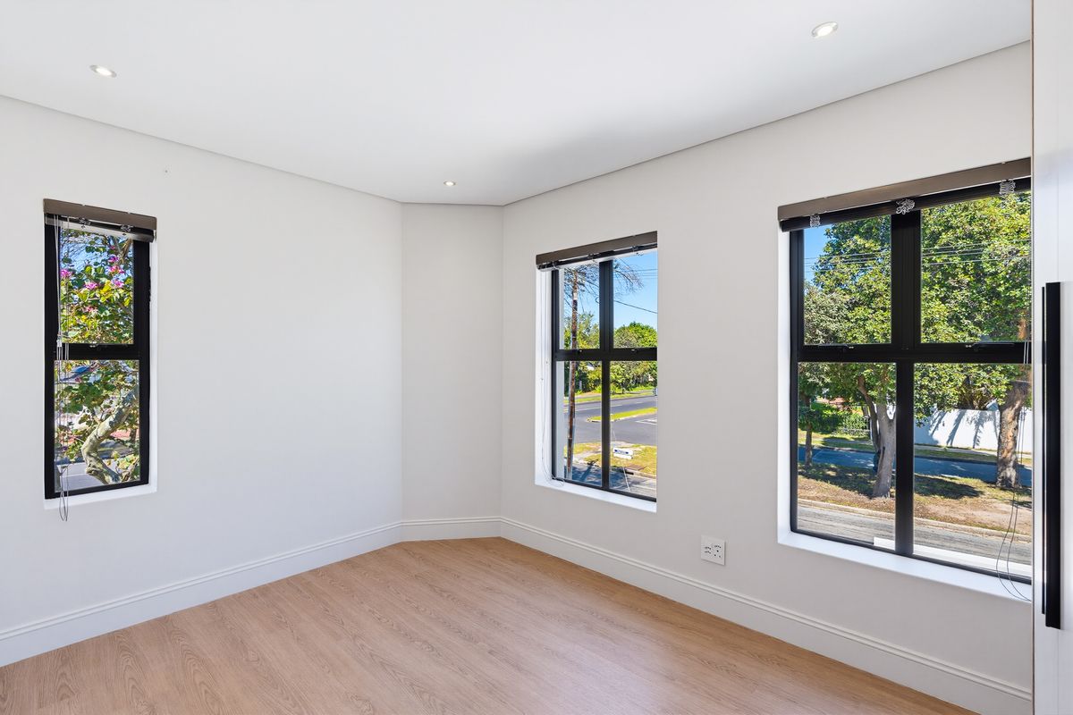 A bright, empty bedroom featuring modern laminate flooring, clean white walls, and large black aluminium-framed windows providing ample natural light.