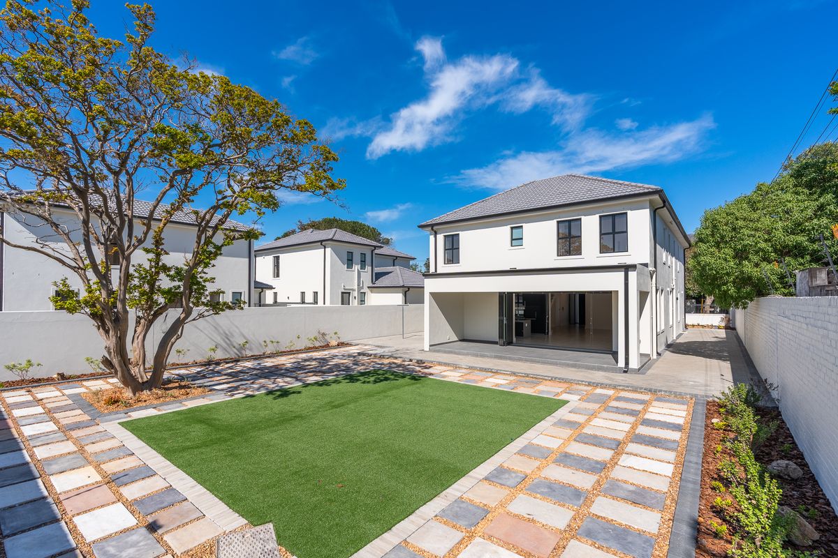 Rear view of a modern two-story house featuring a landscaped garden with artificial turf, paved patio area, and large bi-fold doors leading indoors.