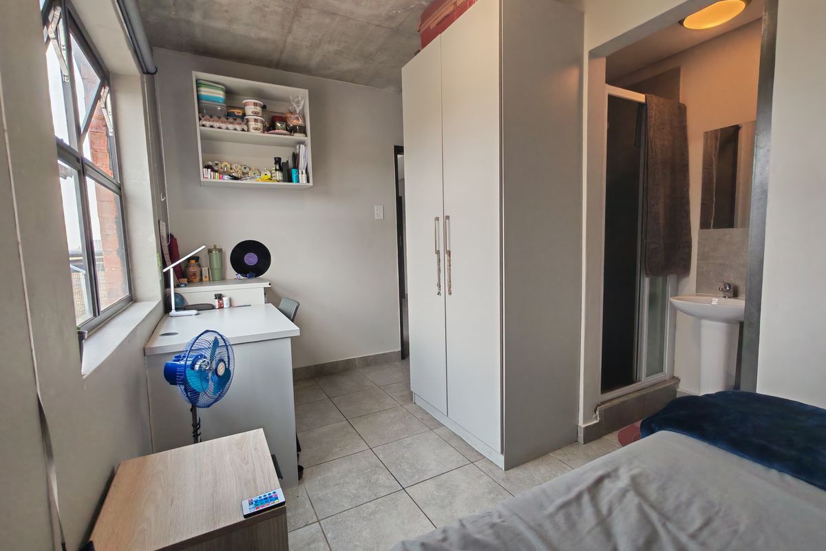 A modern bedroom featuring an exposed concrete ceiling, tiled flooring, built-in wardrobe, and an en-suite bathroom with a pedestal basin.