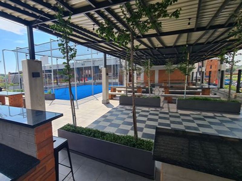 A modern, covered outdoor clubhouse area featuring a built-in bar with granite countertops, tiled flooring, and integrated planters overlooking a sports facility.