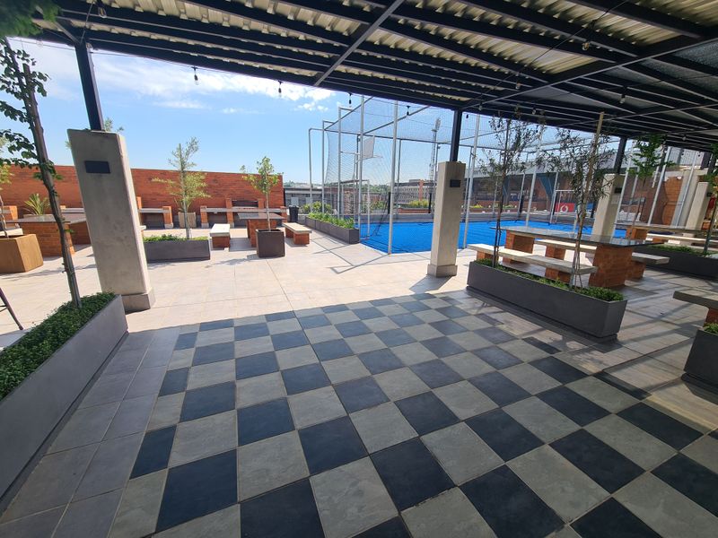 Spacious covered outdoor patio featuring checkered tile flooring, built-in brick seating, and integrated planters, overlooking a private sports facility.