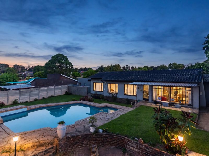 Rear view of the property featuring a well-maintained swimming pool, paved patio area, and established garden under a twilight sky.