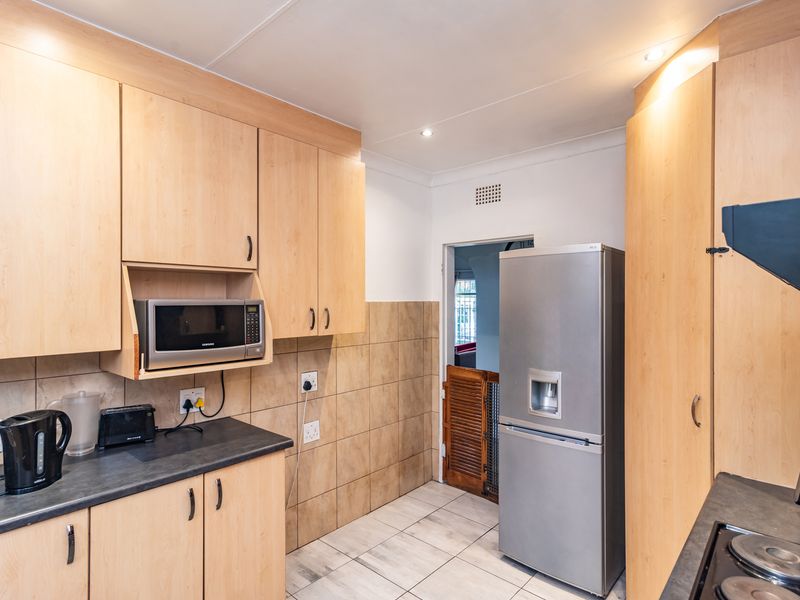 A functional kitchen featuring light wood-finish cabinetry, dark laminate countertops, tiled splashbacks, and tiled flooring with integrated appliances.