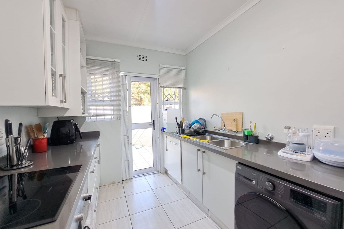 A functional galley-style kitchen featuring white cabinetry, stone countertops, integrated hob and oven, tiled flooring, and direct access to the rear exterior.