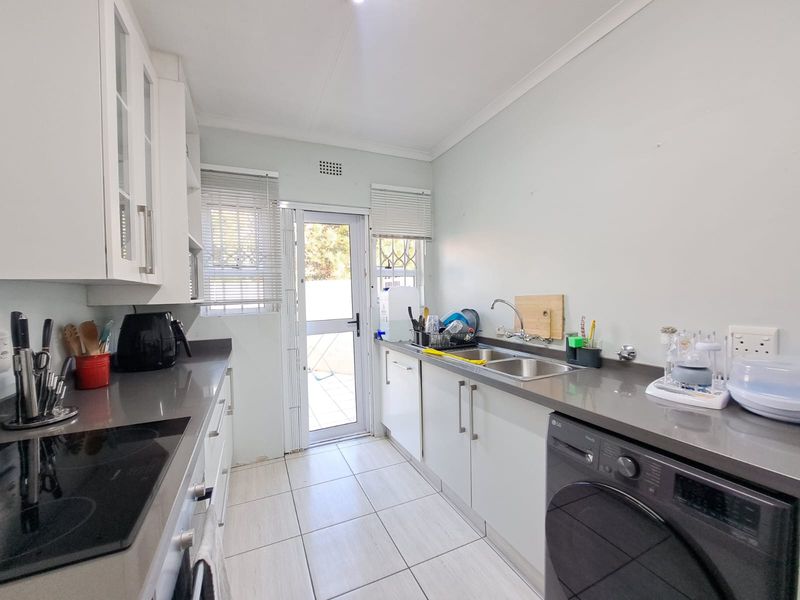 A functional galley-style kitchen featuring white cabinetry, stone countertops, integrated hob and oven, tiled flooring, and direct access to the rear exterior.