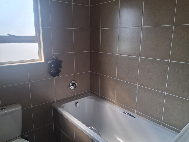 A functional bathroom featuring a bathtub with a mixer tap, a toilet, and neutral wall tiling, complemented by a frosted window for privacy.