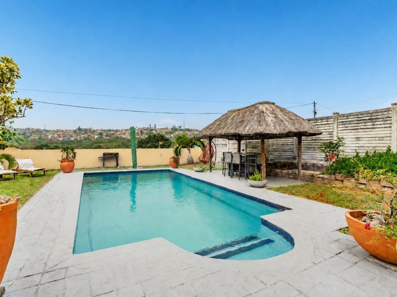 Outdoor pool area featuring a swimming pool, paved surround, and a thatched lapa, set against a boundary wall with distant neighbourhood views.