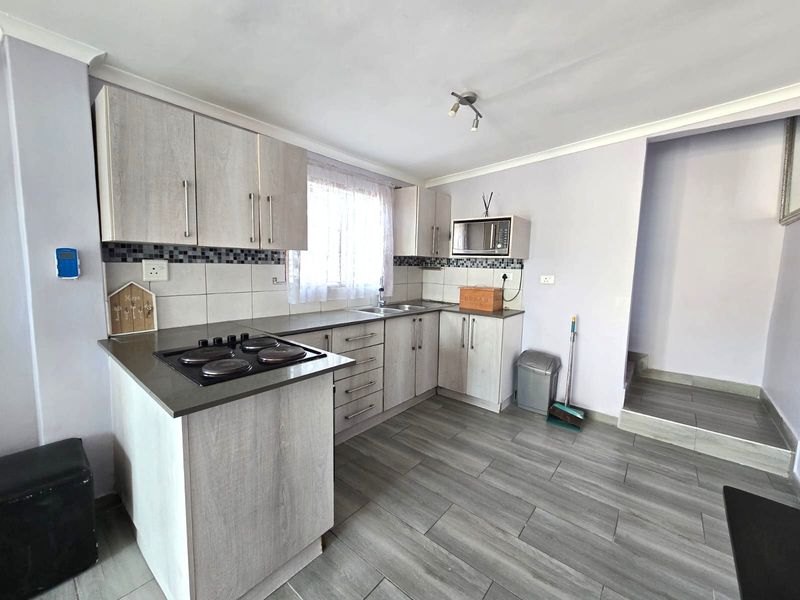 A functional kitchen featuring light wood-grain cabinetry, a built-in electric hob, tiled backsplash, and modern grey floor tiles.