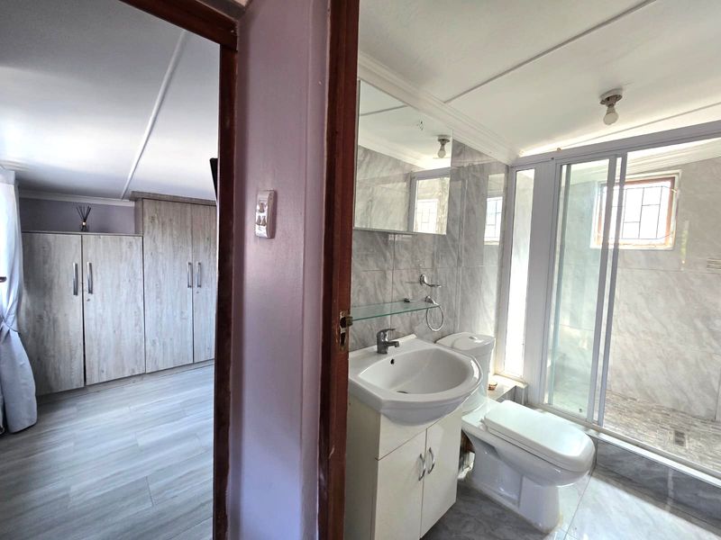 A modern bathroom featuring a vanity basin, toilet, and glass-enclosed shower, with adjacent bedroom storage visible through the doorway.
