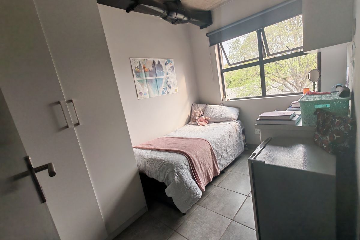 A compact bedroom featuring tiled flooring, a built-in wardrobe, and industrial-style exposed ceiling ducting with natural light from a casement window.