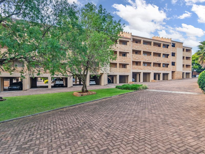 Exterior view of a multi-story apartment complex featuring brick facade, covered parking bays, and a paved driveway with landscaped grounds.