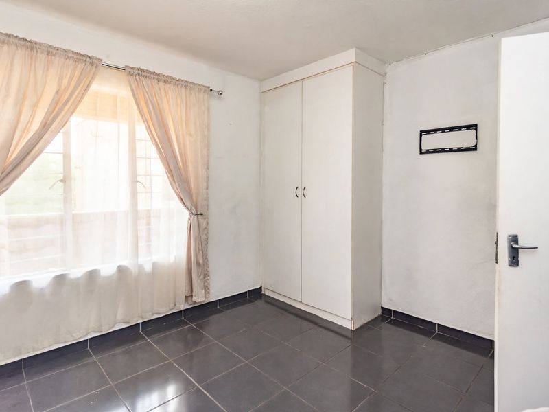 A simple, empty bedroom featuring dark tiled flooring, a built-in wardrobe, and natural light from a window with existing curtain fixtures.