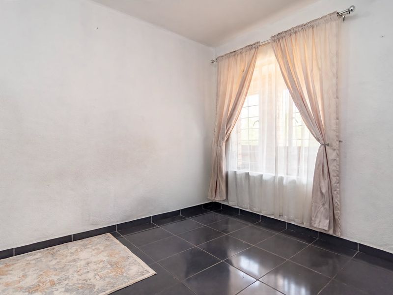 An empty bedroom featuring dark tiled flooring, white walls, and a window fitted with security bars and curtains.