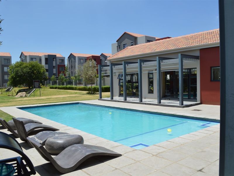 A well-maintained communal swimming pool area featuring a paved deck, pergola structure, and landscaped garden surroundings within a modern apartment complex.