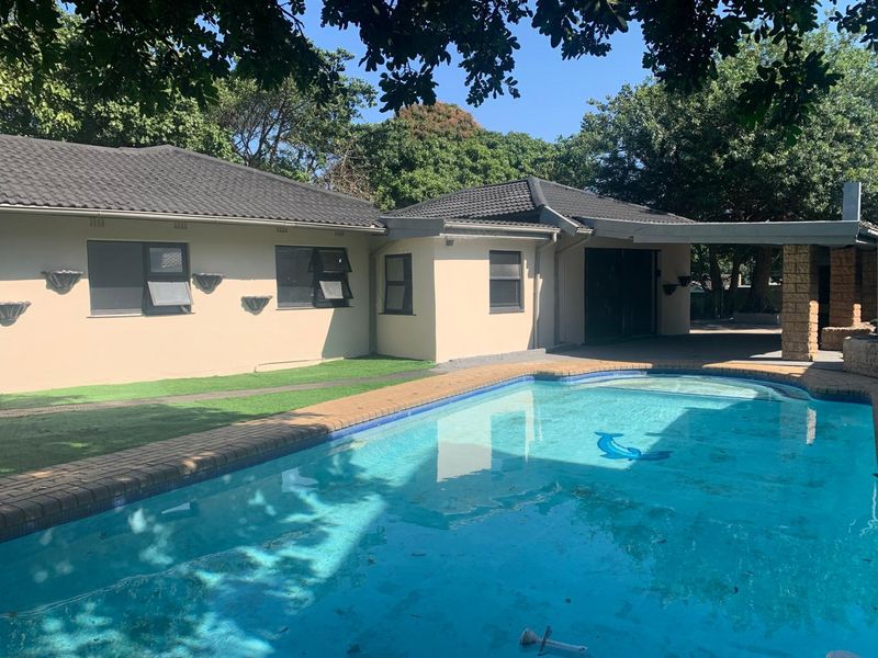 A well-maintained outdoor pool area featuring a swimming pool with brick coping, adjacent to a covered patio and the rear facade of the house.