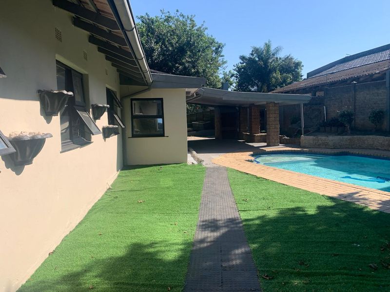 Rear exterior view featuring a swimming pool, covered patio area, brick paving, and artificial lawn, providing a functional outdoor entertainment space.