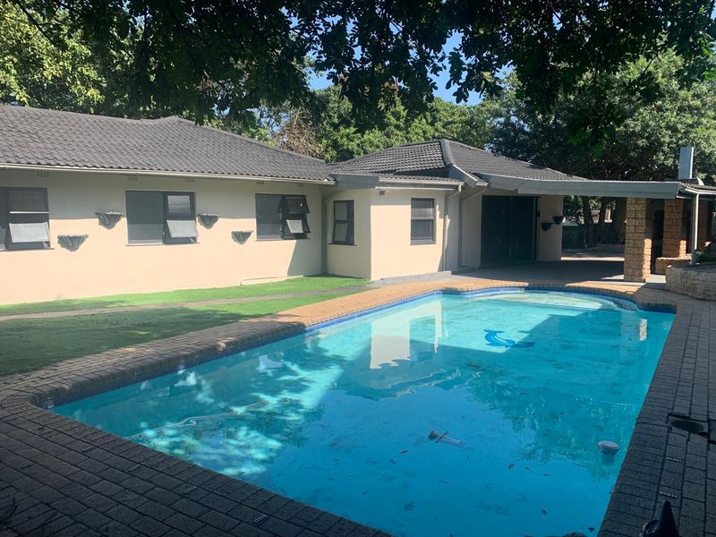 A well-maintained rear garden area featuring a swimming pool, brick-paved surround, and a covered patio with a built-in braai.