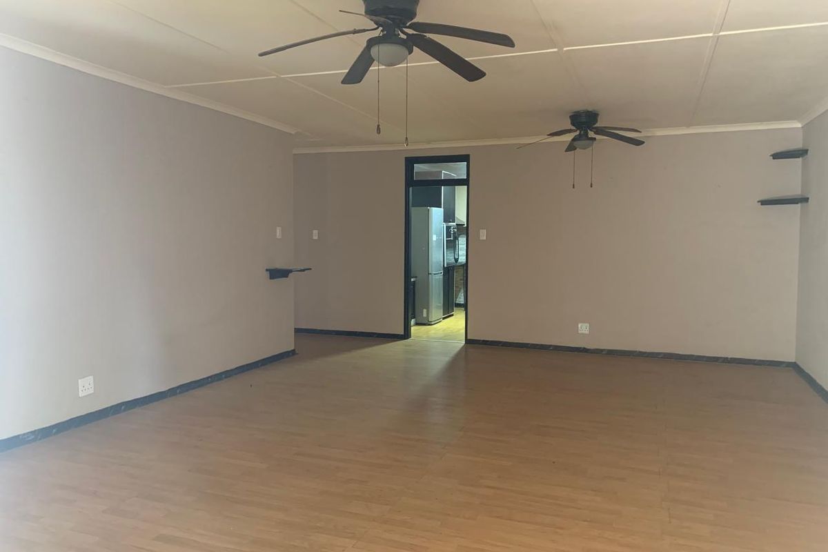 A spacious, empty living area featuring laminate flooring, dual ceiling fans, and neutral wall tones, with a direct view into the adjacent kitchen.