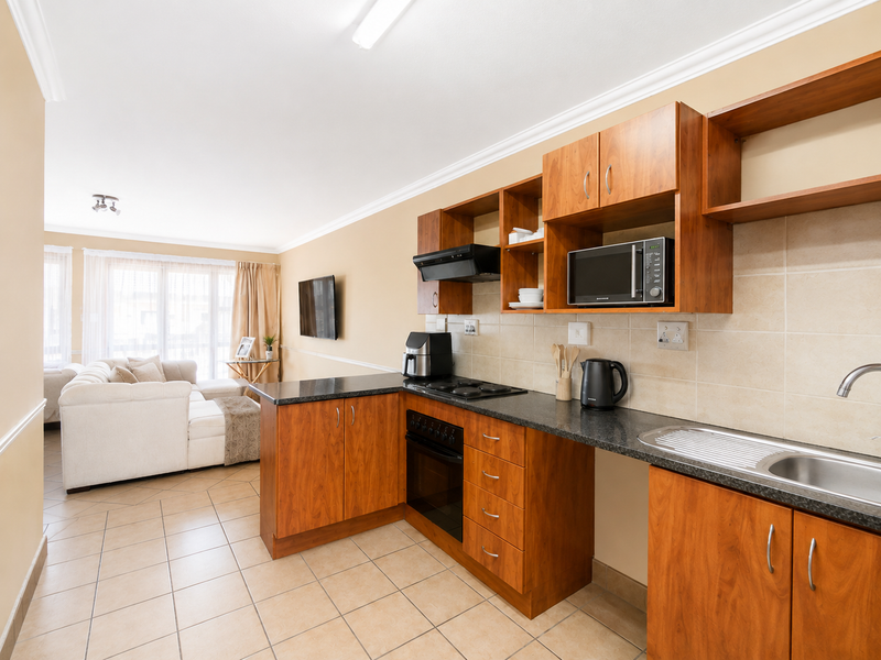 Open plan kitchen featuring wood-finish cabinetry, dark countertops, and integrated appliances, flowing into a bright living area with tiled floors.