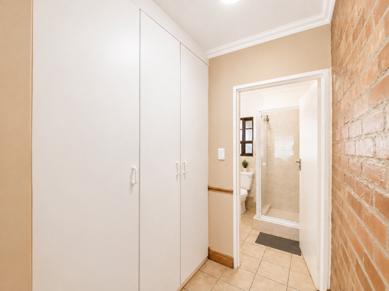 A clean hallway featuring built-in white cupboards, an exposed brick feature wall, and tiled flooring, leading directly into a bathroom.